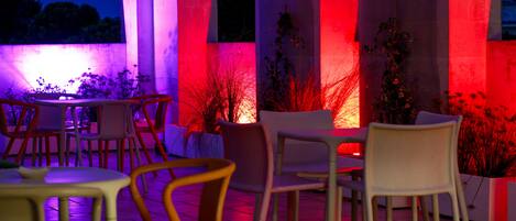 Un espacio al aire libre con mesas y sillas, iluminado con luces rojas y púrpuras, rodeado de columnas y plantas.