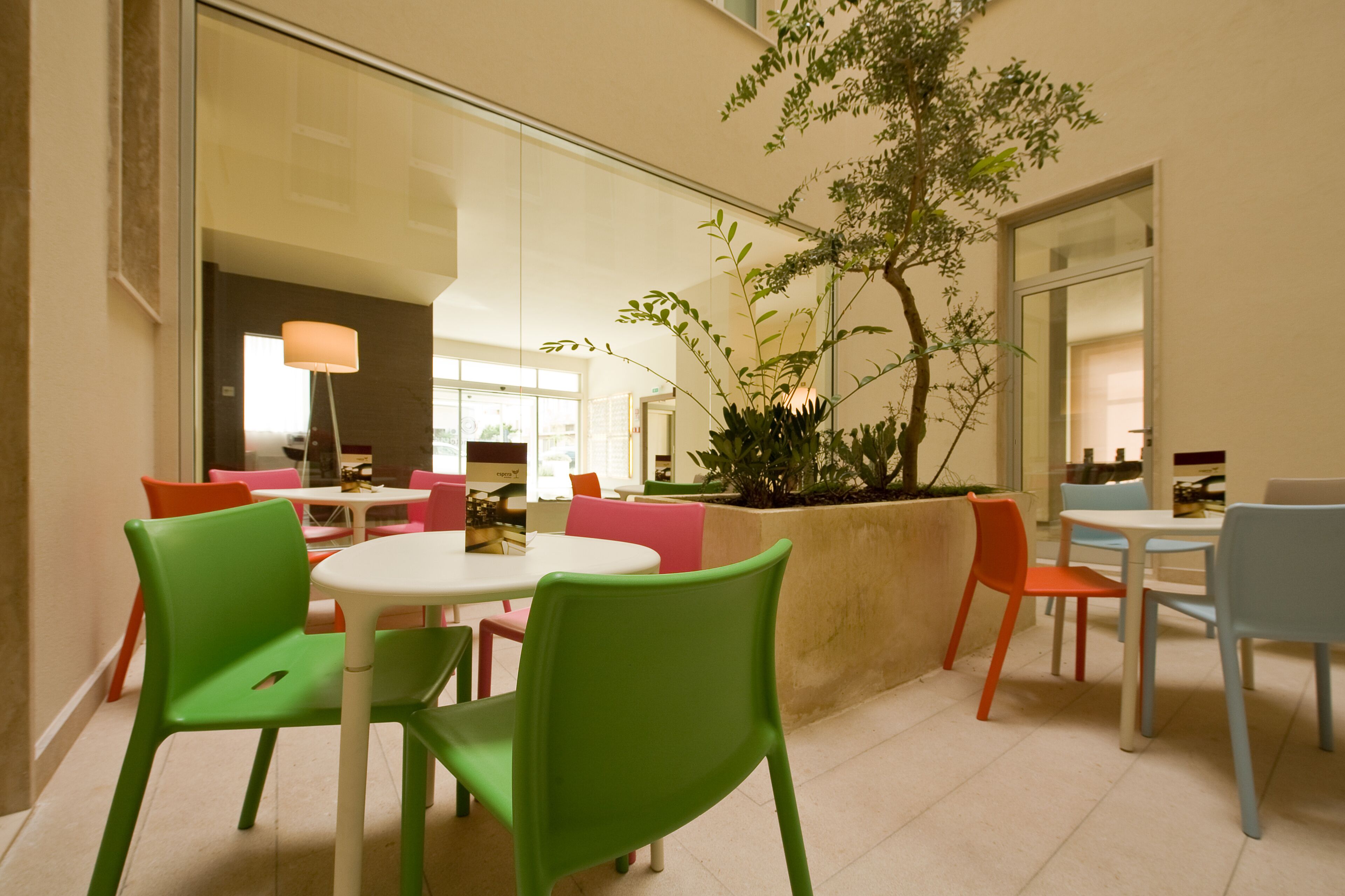 Un comedor con sillas y mesas coloridas, un ventanal grande y una planta en maceta.