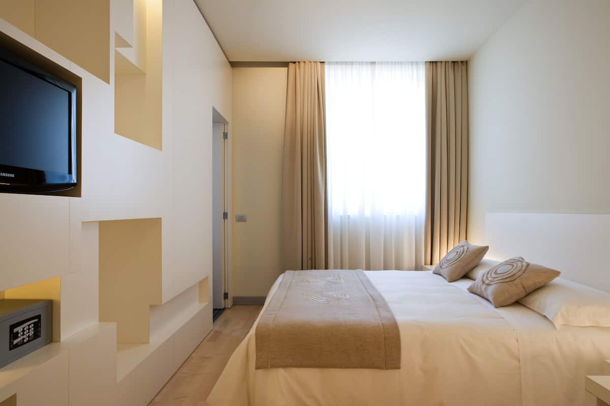 Ett modernt hotellrum med en stor säng, en platt-TV som är monterad på väggen och en kontrollpanel för rummets olika funktioner.
