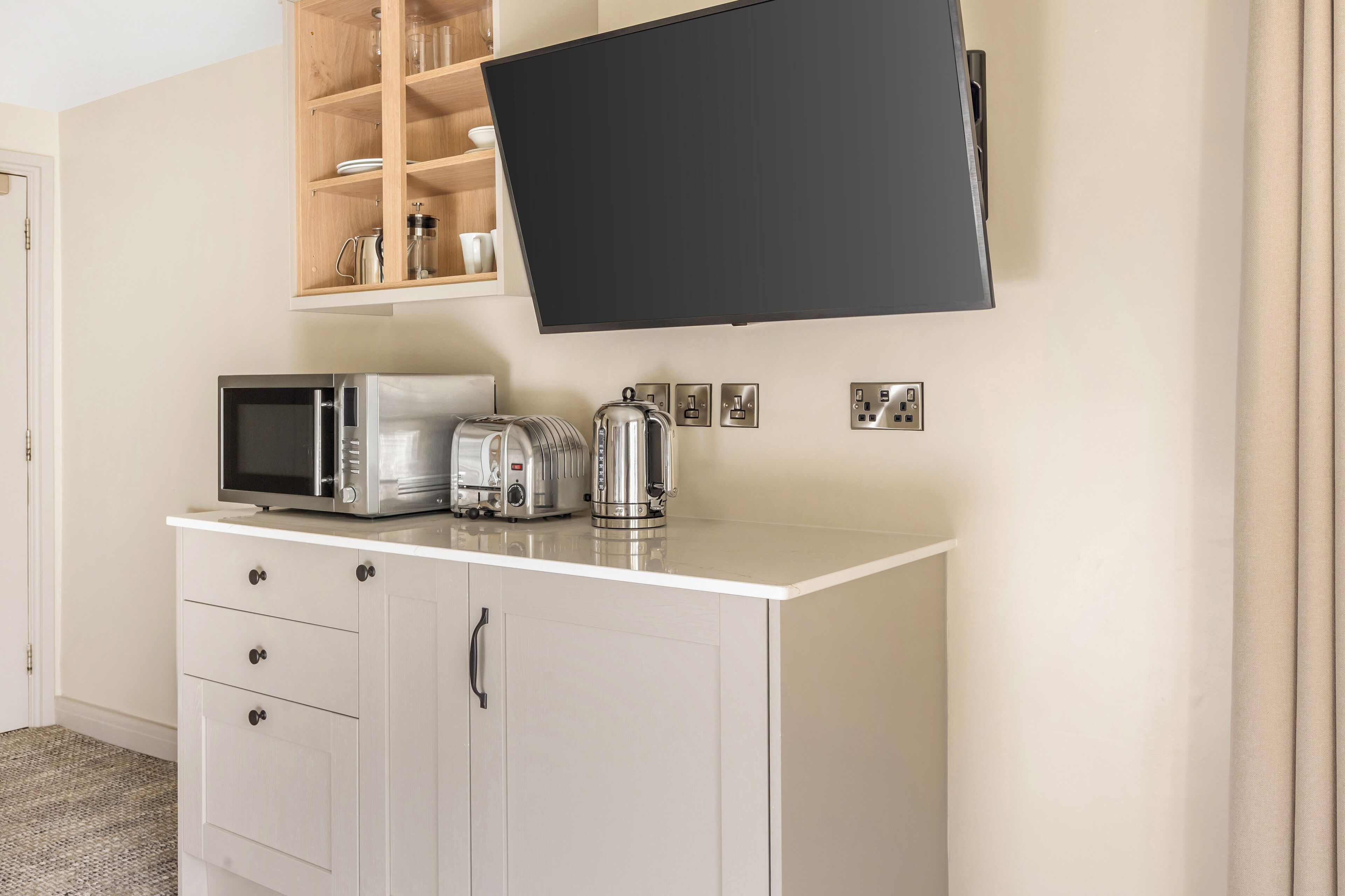 Un angolo cottura con forno a microonde, tostapane e bollitore su un ripiano bianco, un armadietto in legno sopra e una TV a schermo piatto montata a parete.