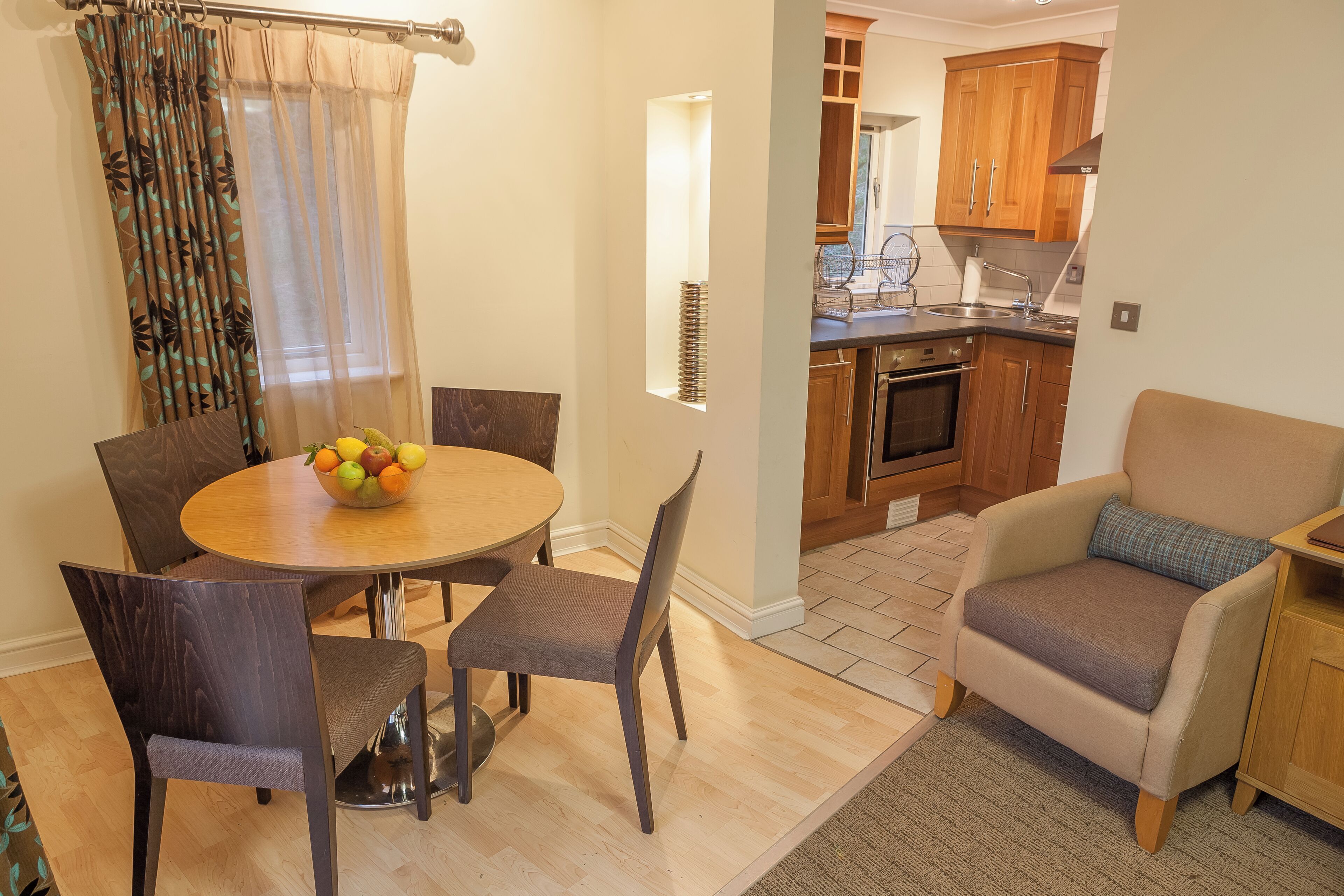 A dining area with a round table, chairs, and a fruit bowl. There is a kitchen visible in the background with wooden cabinets and a stove.