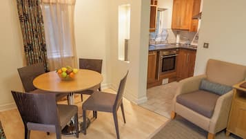 A dining area with a round table, chairs, and a fruit bowl. There is a kitchen visible in the background with wooden cabinets and a stove.