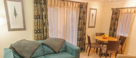 A living room with a teal sofa, patterned cushions, a dining table with chairs, and a fruit bowl.
