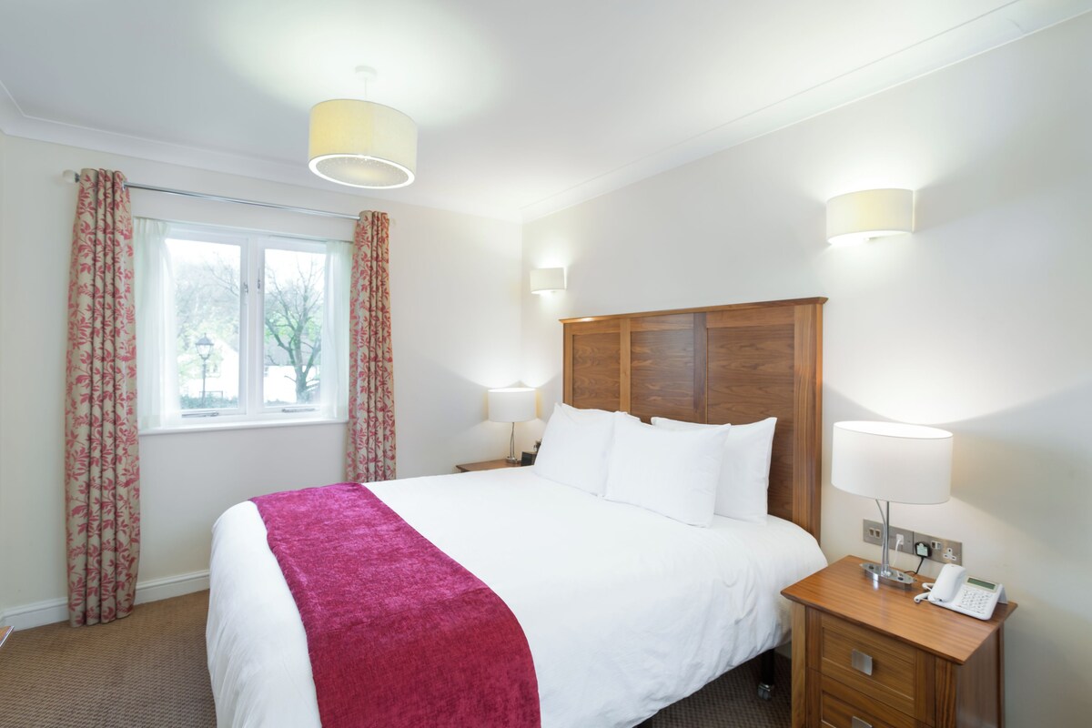 A hotel room with a large bed, two bedside lamps, a wooden nightstand, and a window with curtains.