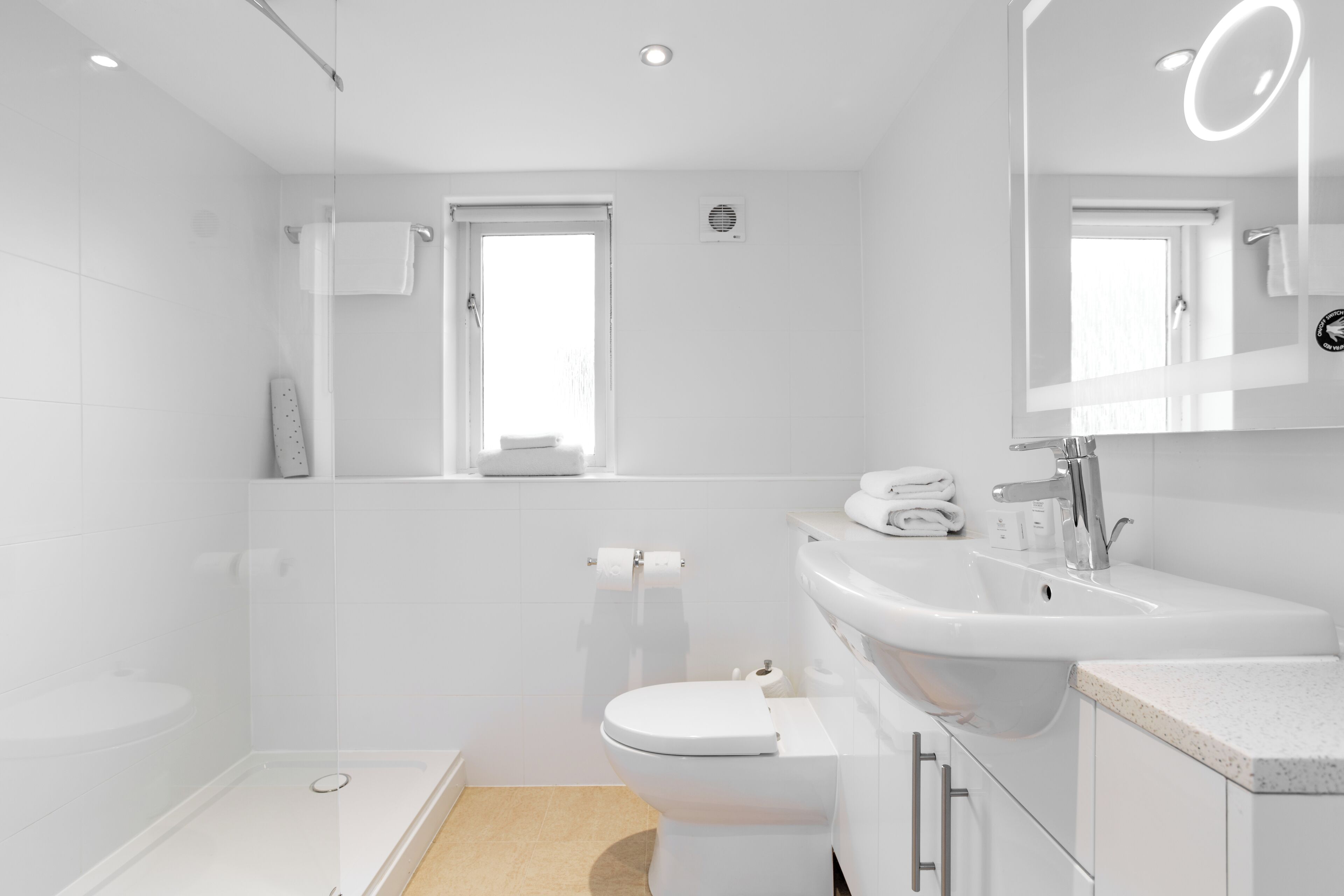 Une salle de bain moderne avec une douche à l’italienne en verre, des toilettes blanches, un lavabo avec un miroir et une fenêtre laissant entrer la lumière naturelle.