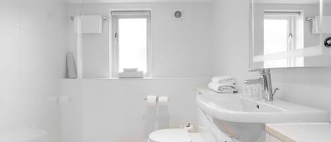 Une salle de bain moderne avec une douche à l’italienne en verre, des toilettes blanches, un lavabo avec un miroir et une fenêtre laissant entrer la lumière naturelle.