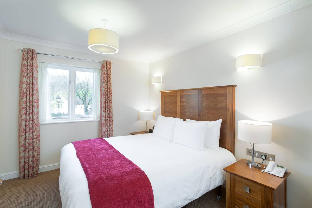 A hotel room with a large bed, two bedside lamps, a wooden nightstand, and a window with curtains.