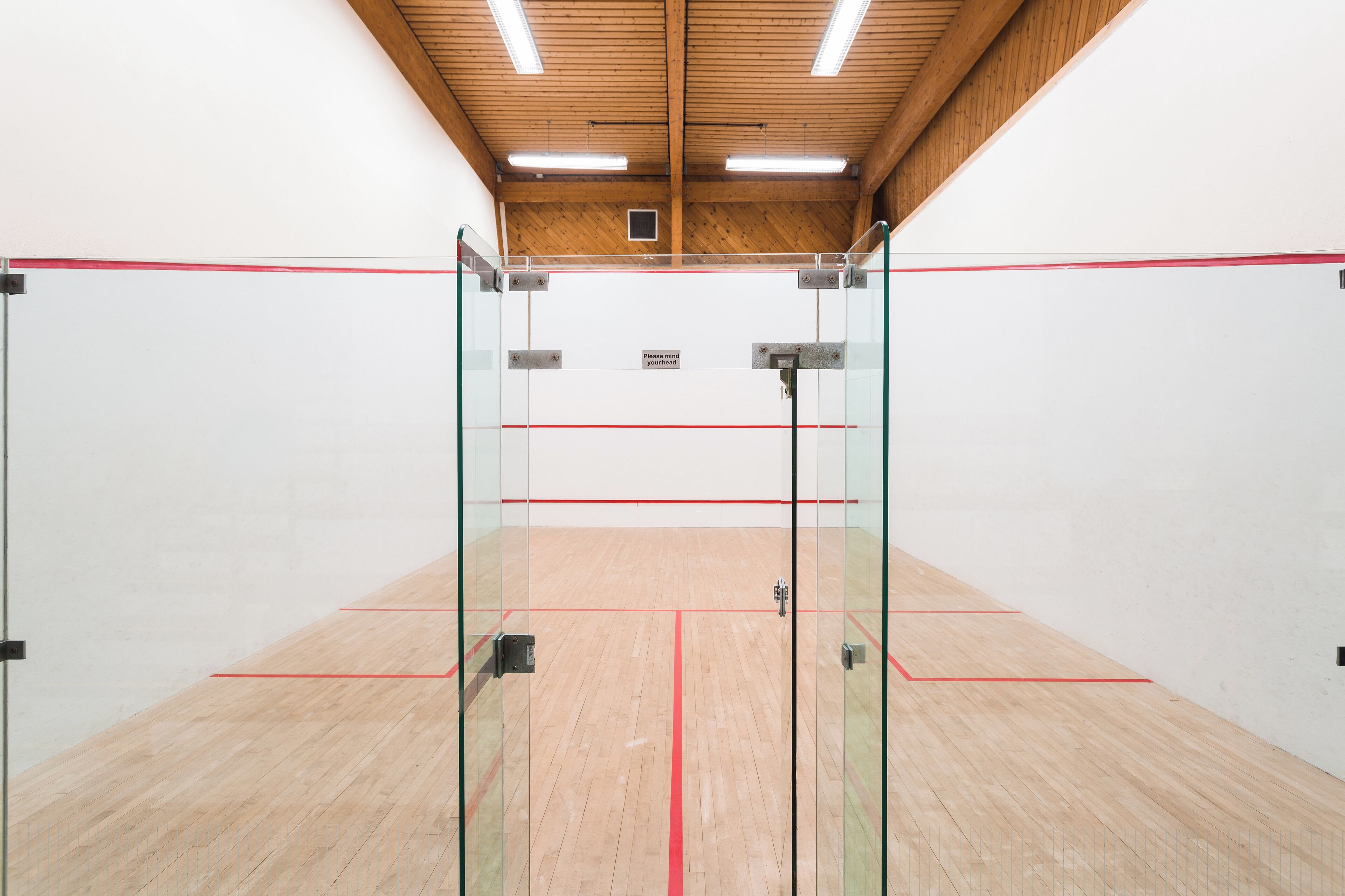 Ein Squashplatz mit Glaswänden, Holzboden und Deckenleuchten.