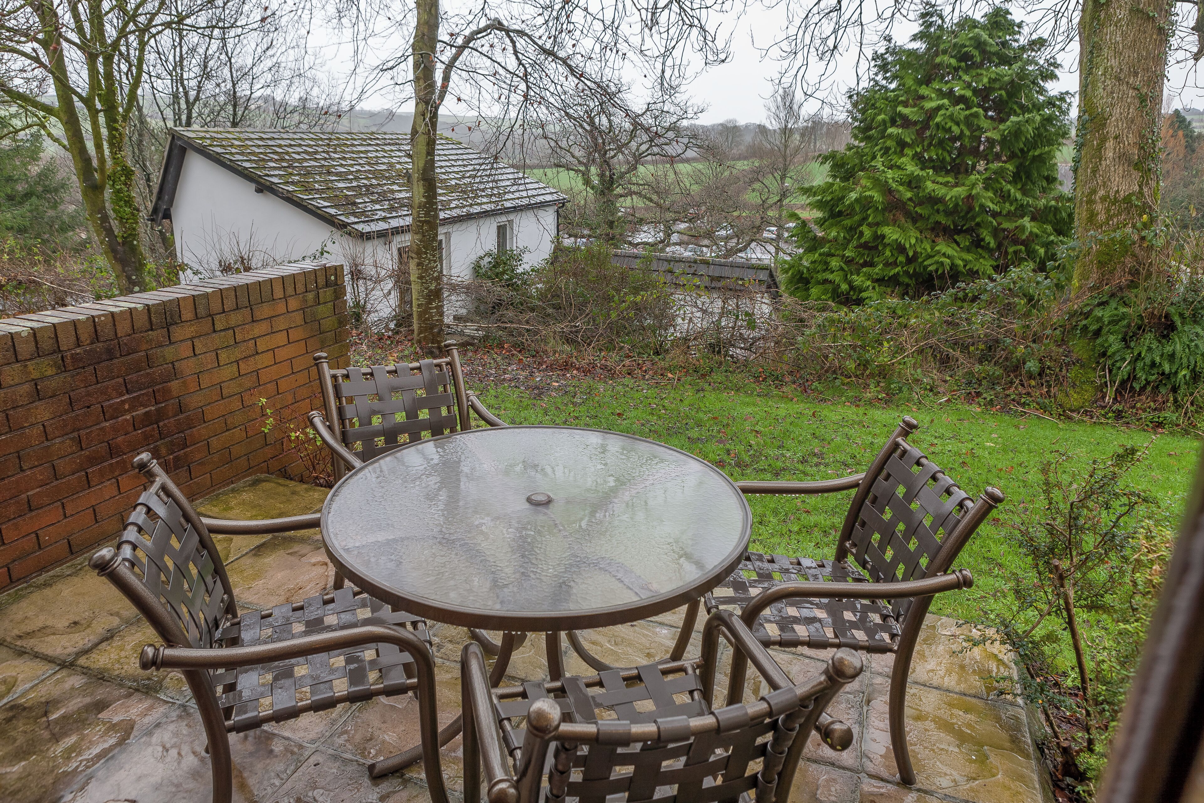 A patio with a round glass table and wicker chairs, surrounded by a brick wall and a grassy area with trees.