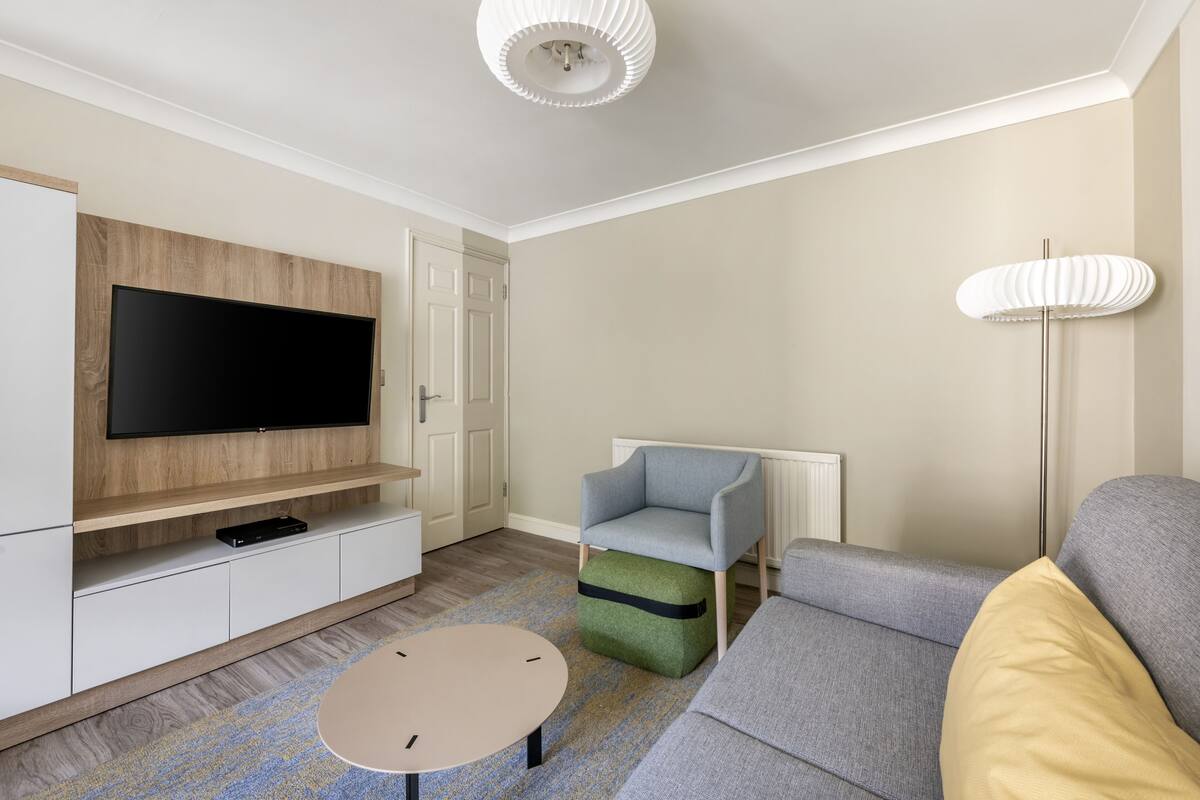 A modern living room with a flat-screen TV, a grey sofa, a blue armchair, and a small round coffee table.