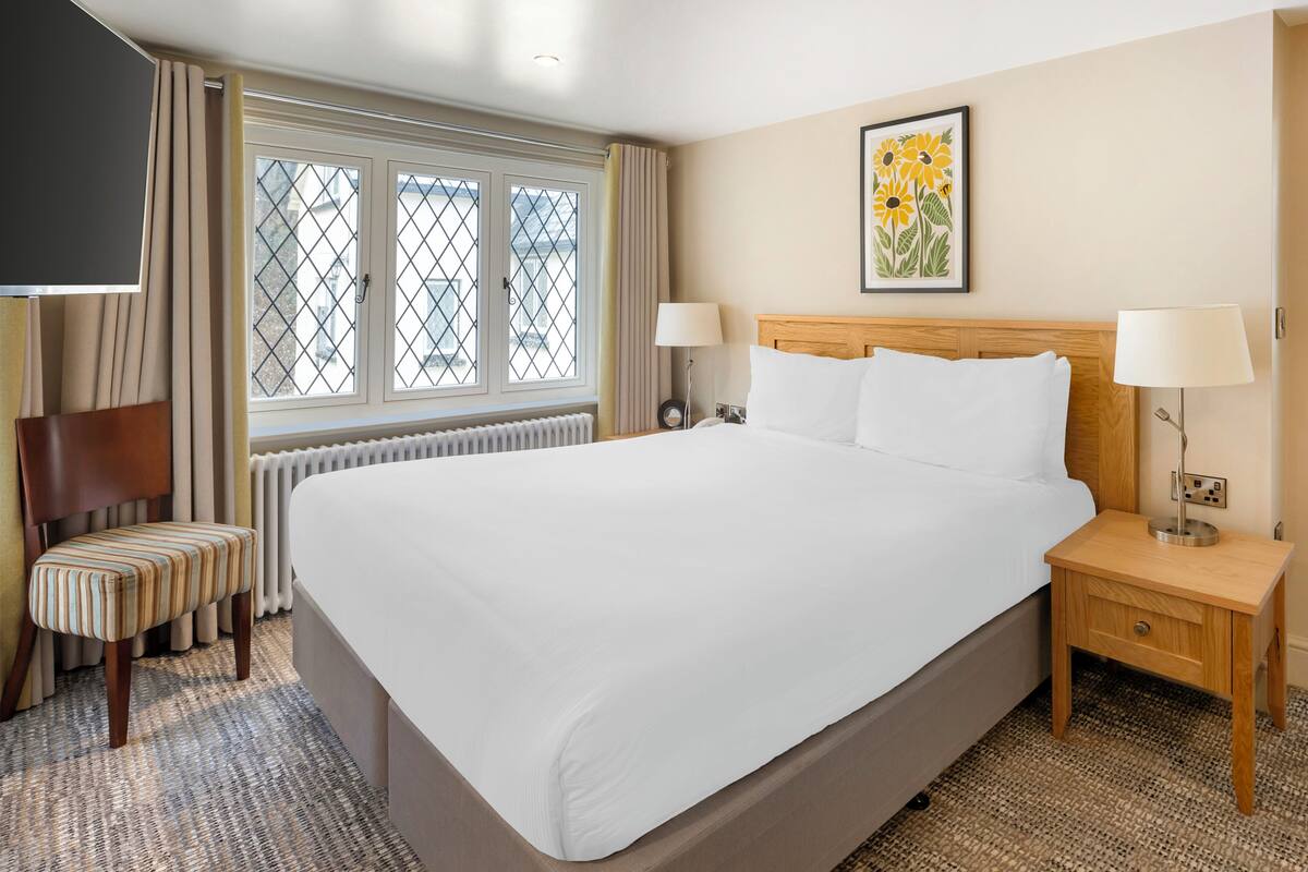 A hotel room with a large bed, two bedside tables, a chair, a window with curtains, and a painting on the wall.