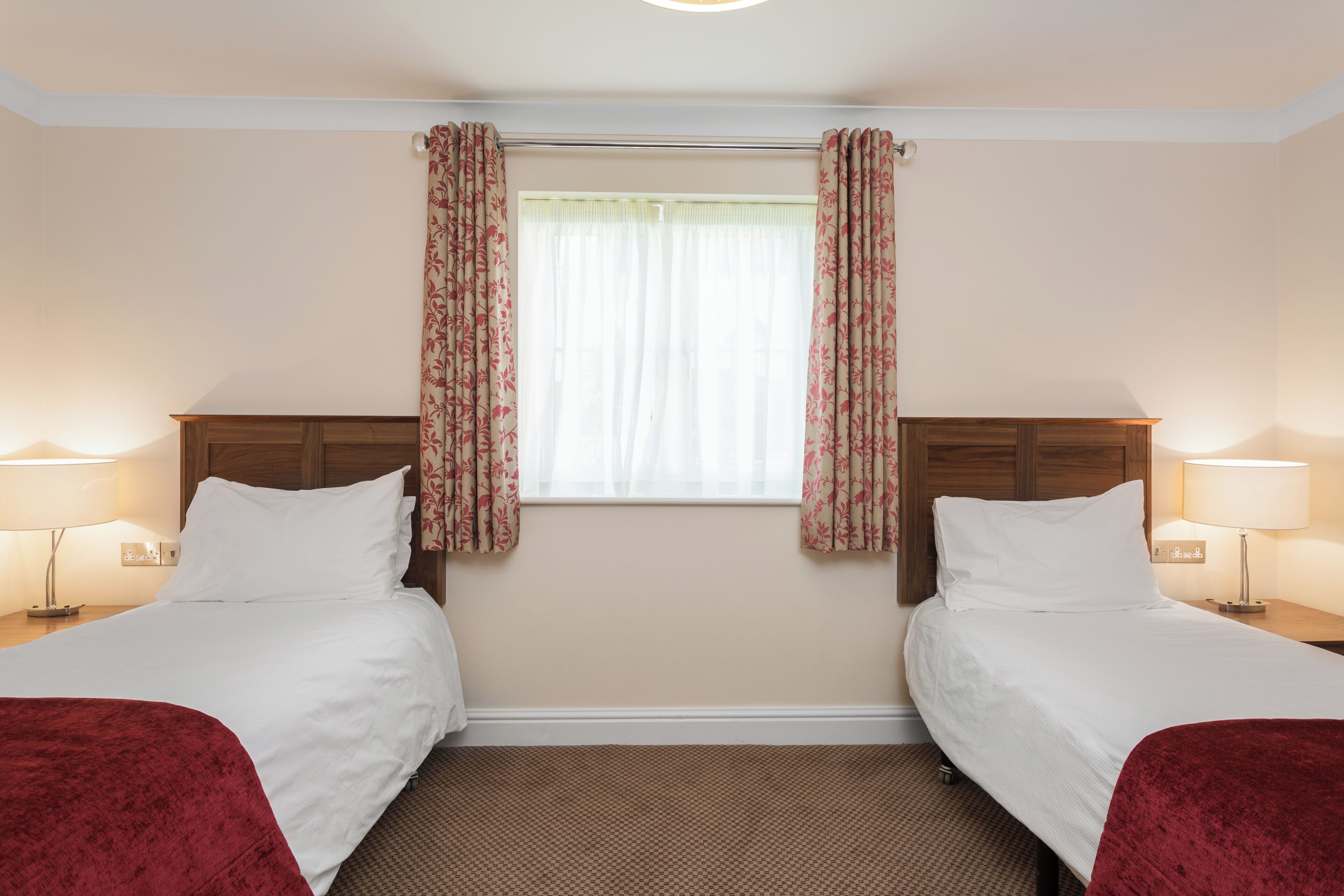 A hotel room with two beds, a window with floral curtains, and bedside lamps.