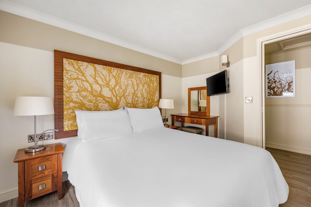 A hotel room with a large bed, two bedside tables with lamps, a flat-screen TV mounted on the wall, and a wooden headboard with a tree pattern.