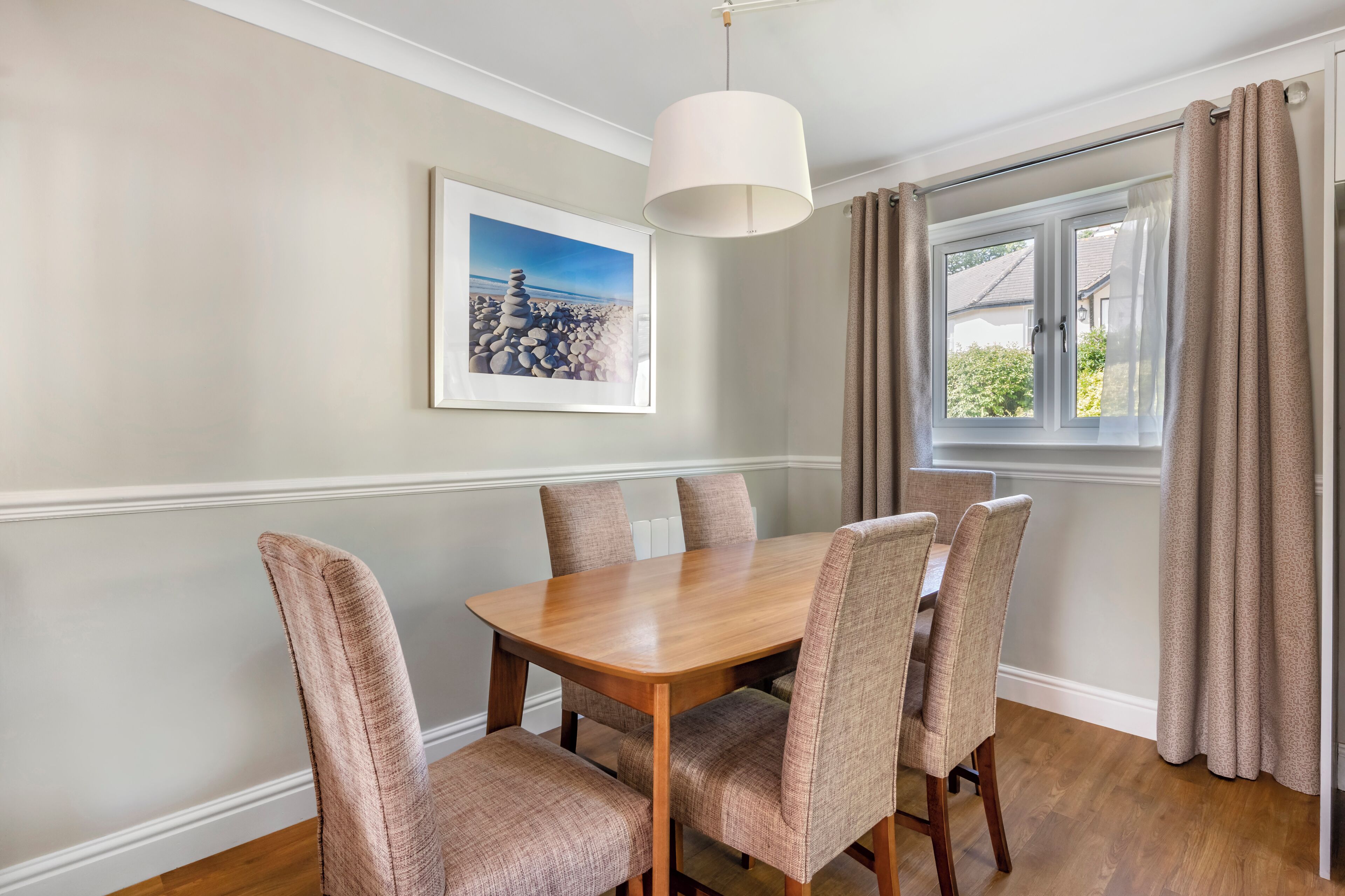 A dining area with a wooden table and cushioned chairs, a pendant light, a framed picture on the wall, and a window with curtains.