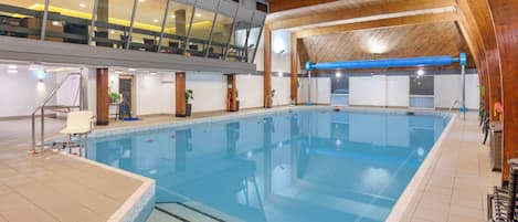 An indoor swimming pool with a tiled floor, a wooden ceiling, and a seating area with chairs and tables.