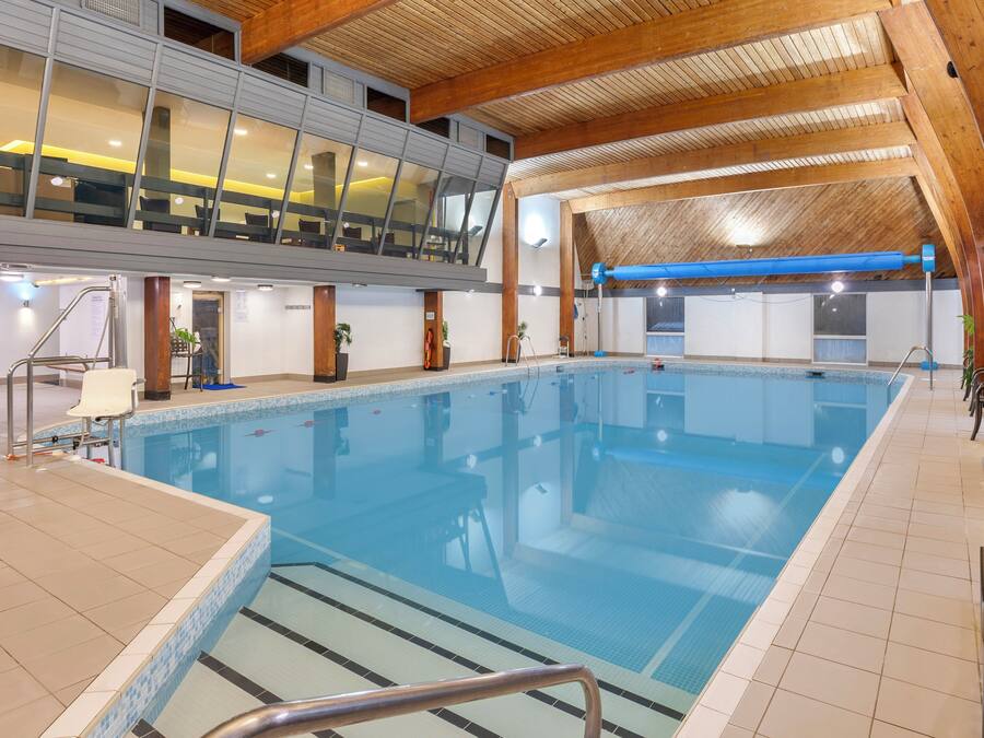 Piscina interna com chão em mosaico, teto de madeira e área de estar com cadeiras e mesas.