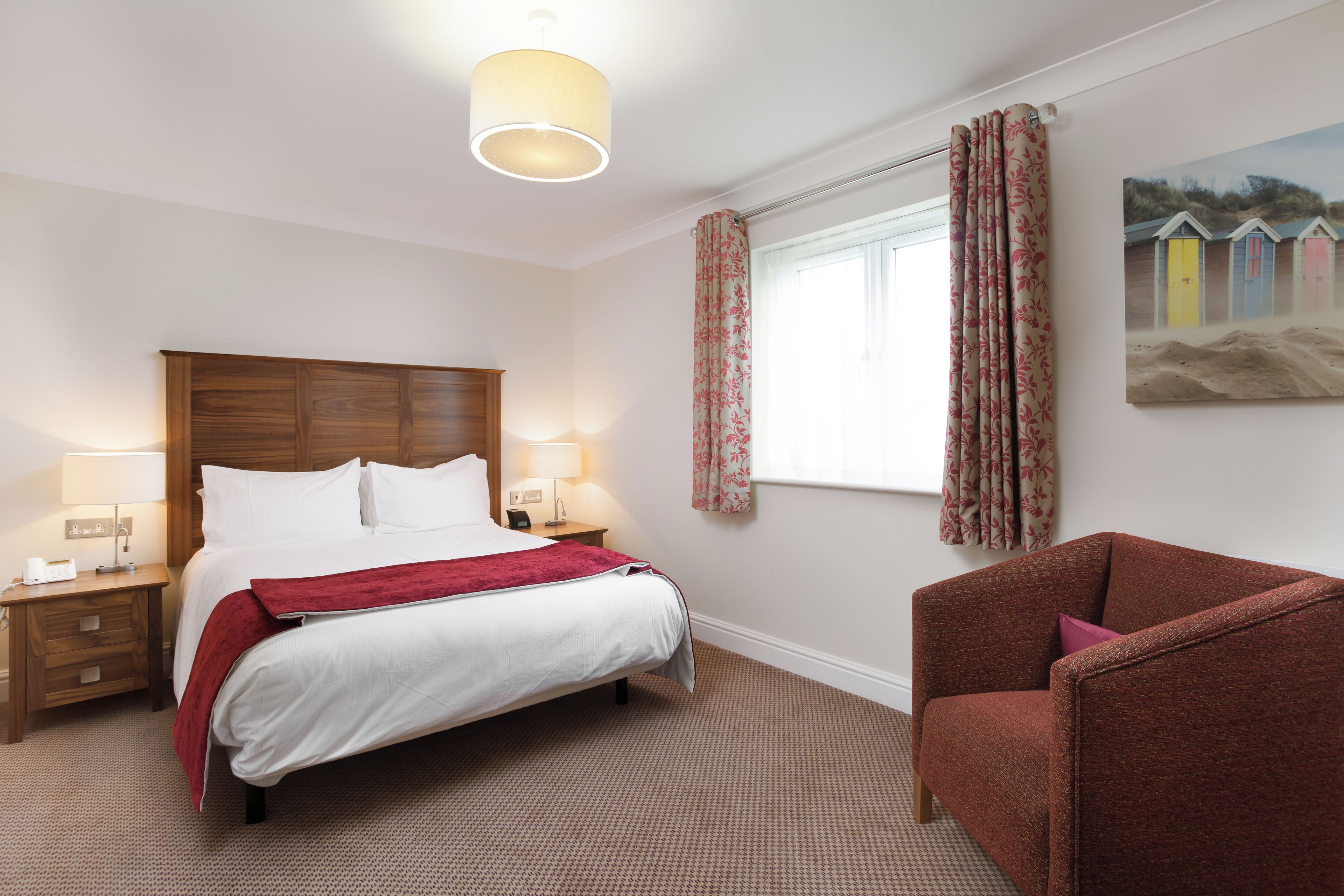 A hotel room with a bed, bedside tables, a chair, a window with curtains, and a wall-mounted painting.