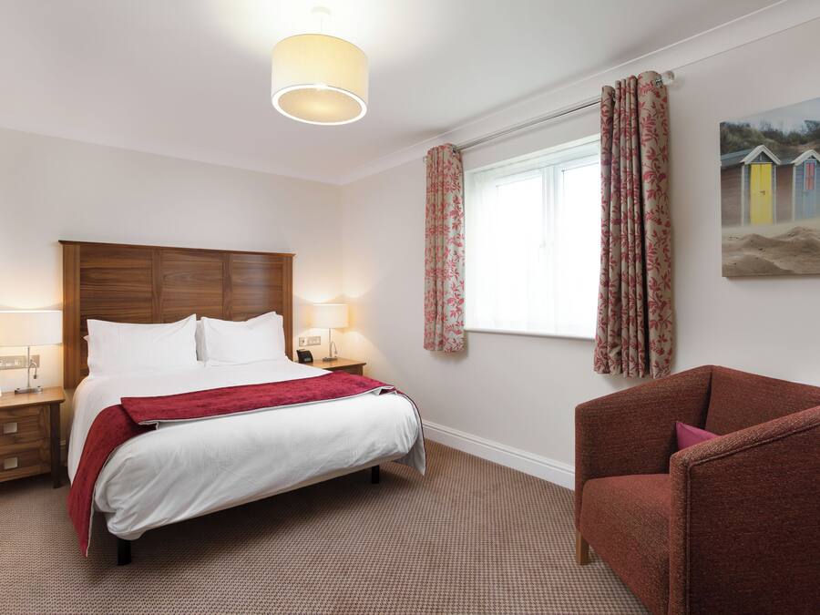 A hotel room with a bed, bedside tables, a chair, a window with curtains, and a wall-mounted painting.