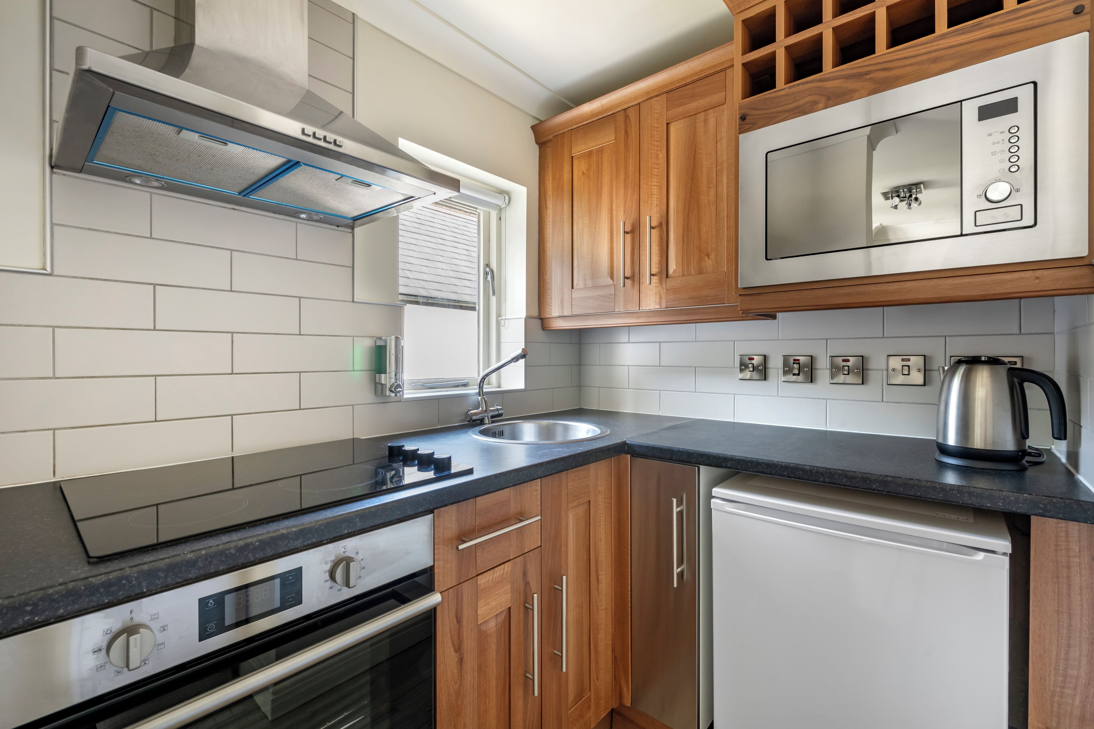 A modern kitchen with wooden cabinets, a stainless steel range hood, a built-in microwave, and a kettle on the countertop.