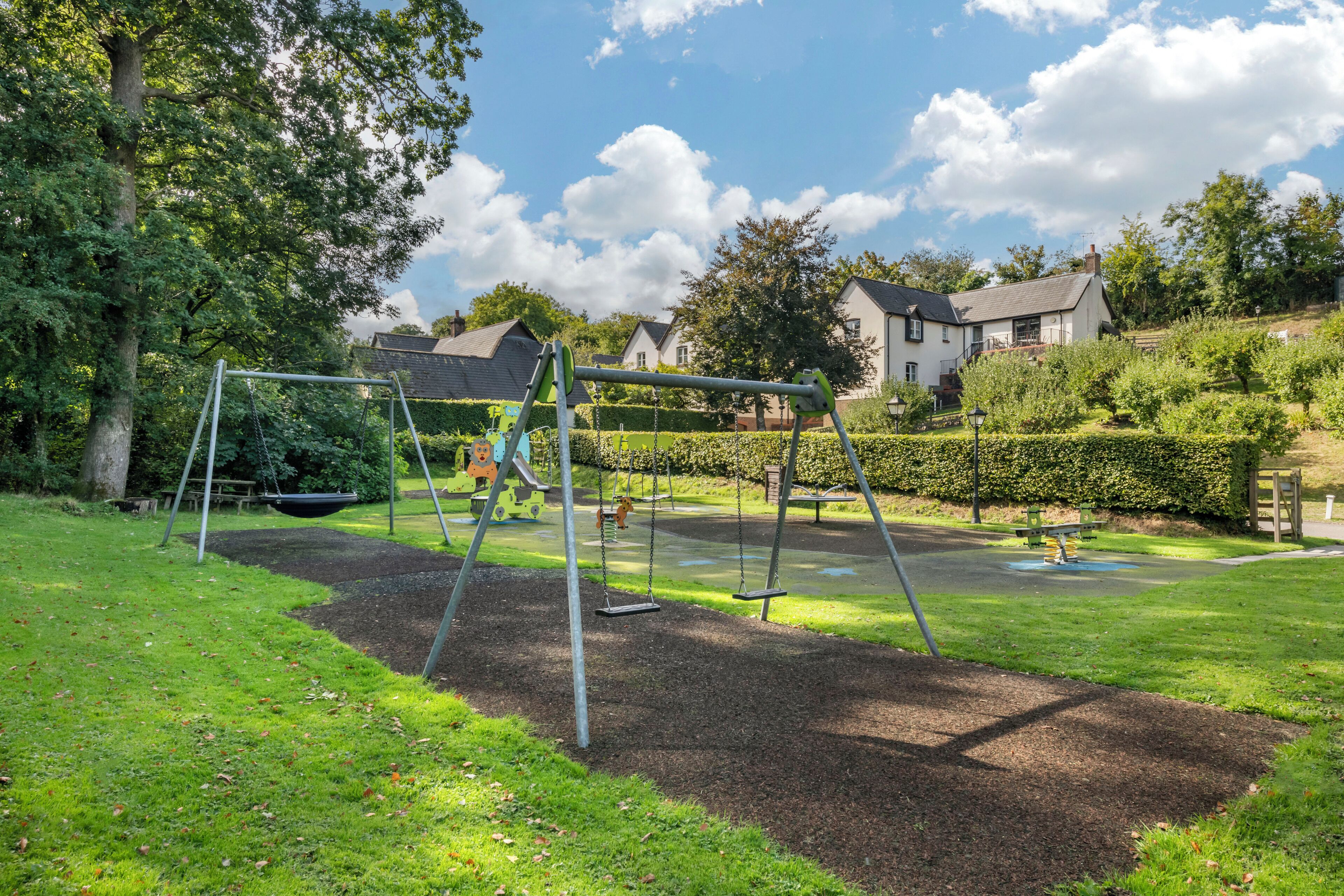 Un parque infantil con columpios y tobogán, rodeado de césped cuidado y árboles.