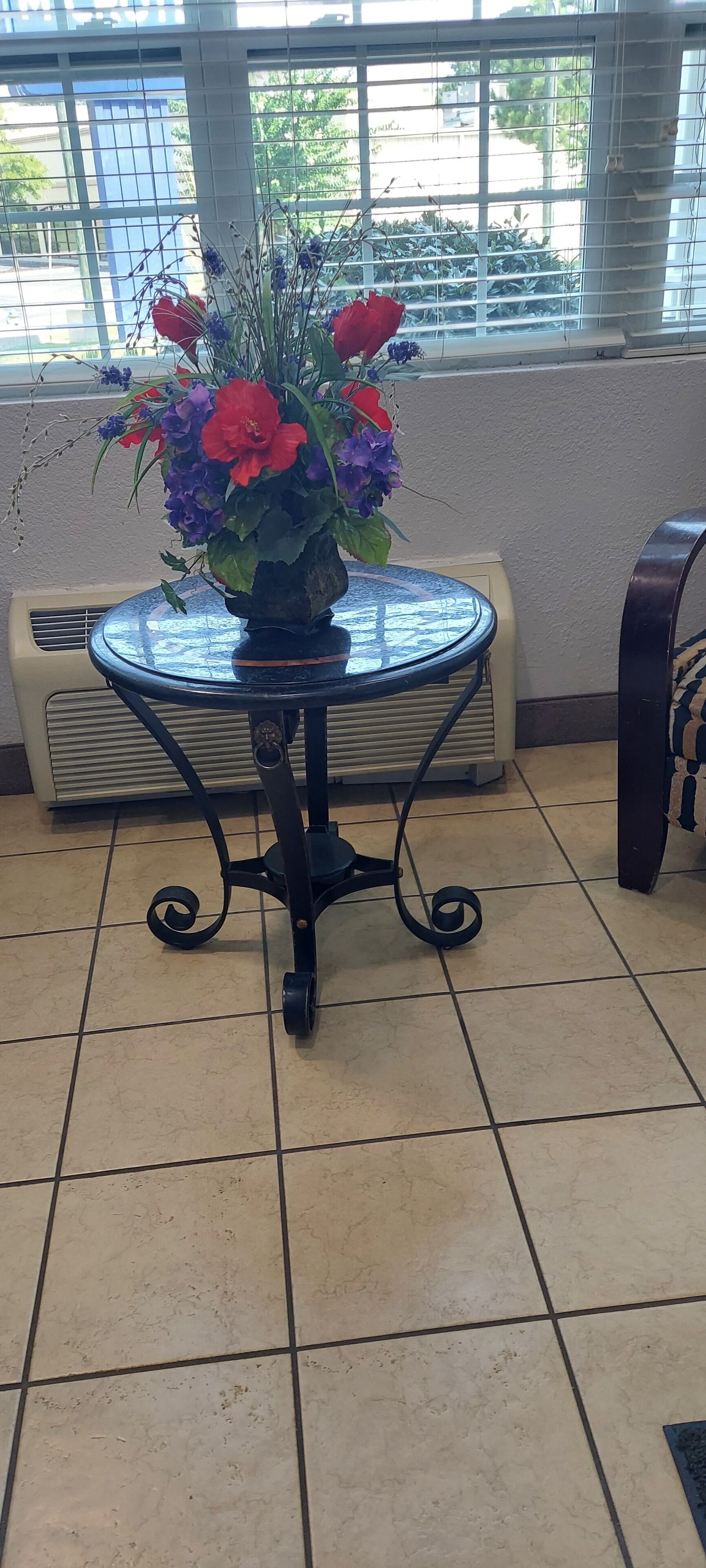 A small round table with a floral arrangement in a room with tiled flooring and a window with blinds.
