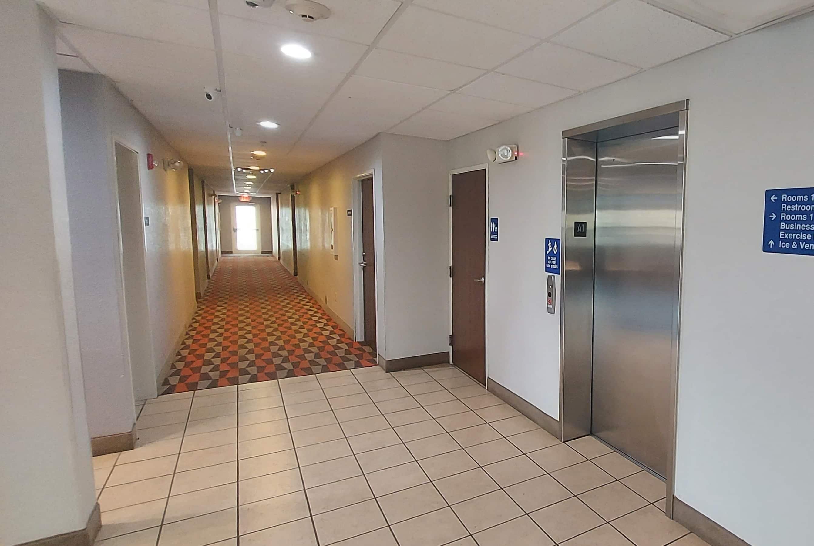 A hotel corridor with a patterned carpet, a central elevator, and directional signs.