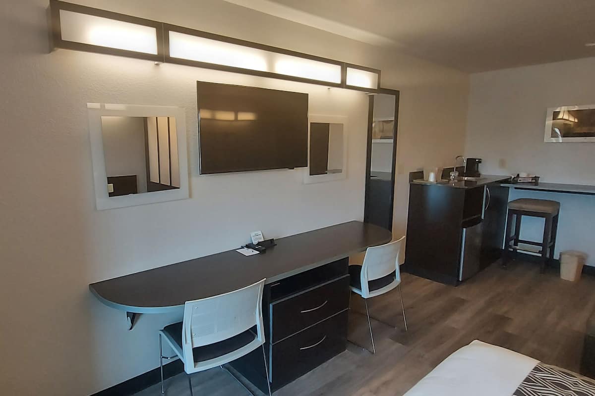 A modern hotel room with a dark wooden desk, white chairs, a large mirror, and a small kitchenette.