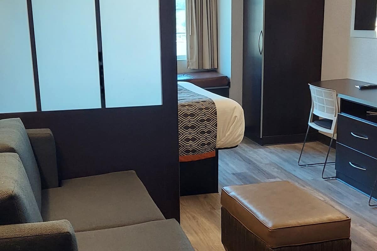 A modern hotel room with a sectional sofa, a small ottoman, a desk with a chair, a wardrobe, and a bed with patterned bedding.