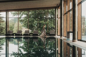 A modern indoor pool area with large windows, wooden columns, and lounge chairs. - Wellnesshotel Eibl-Brunner (Frauenau)