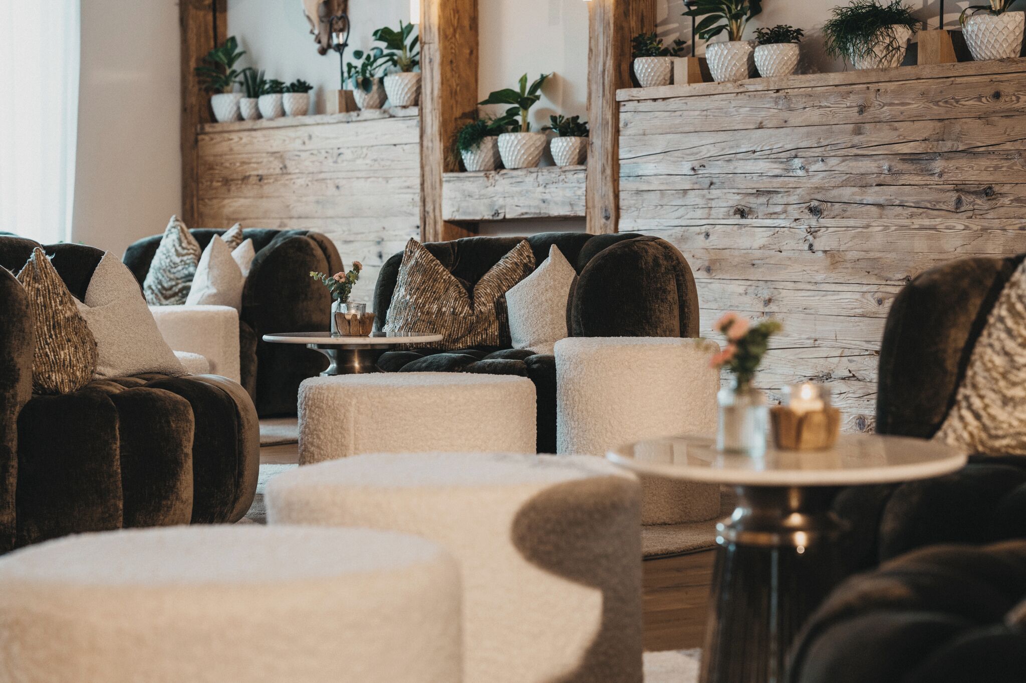 A cozy seating area with plush armchairs and a round table, set against a rustic wooden wall with potted plants.