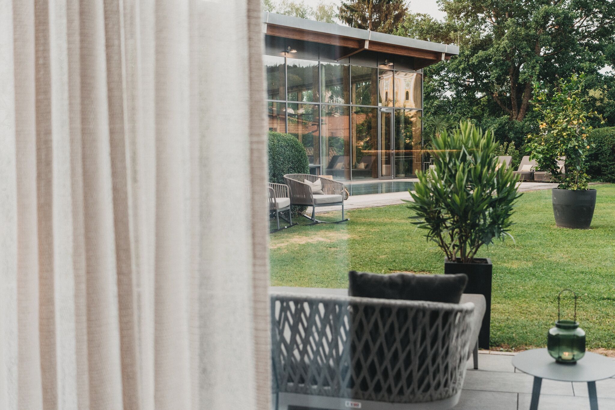 A modern outdoor seating area with a glass wall, a potted plant, and a small table with a lantern.