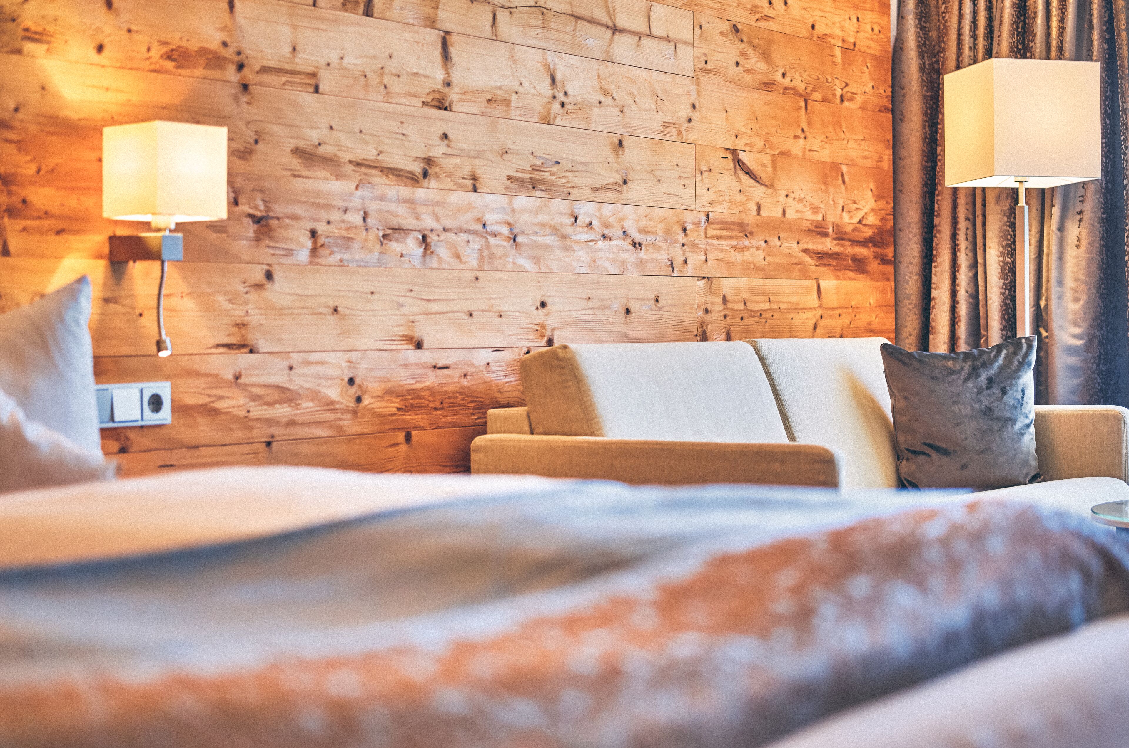 A hotel room with a wooden wall, a bed with a white duvet, a beige sofa, and two wall-mounted lamps.