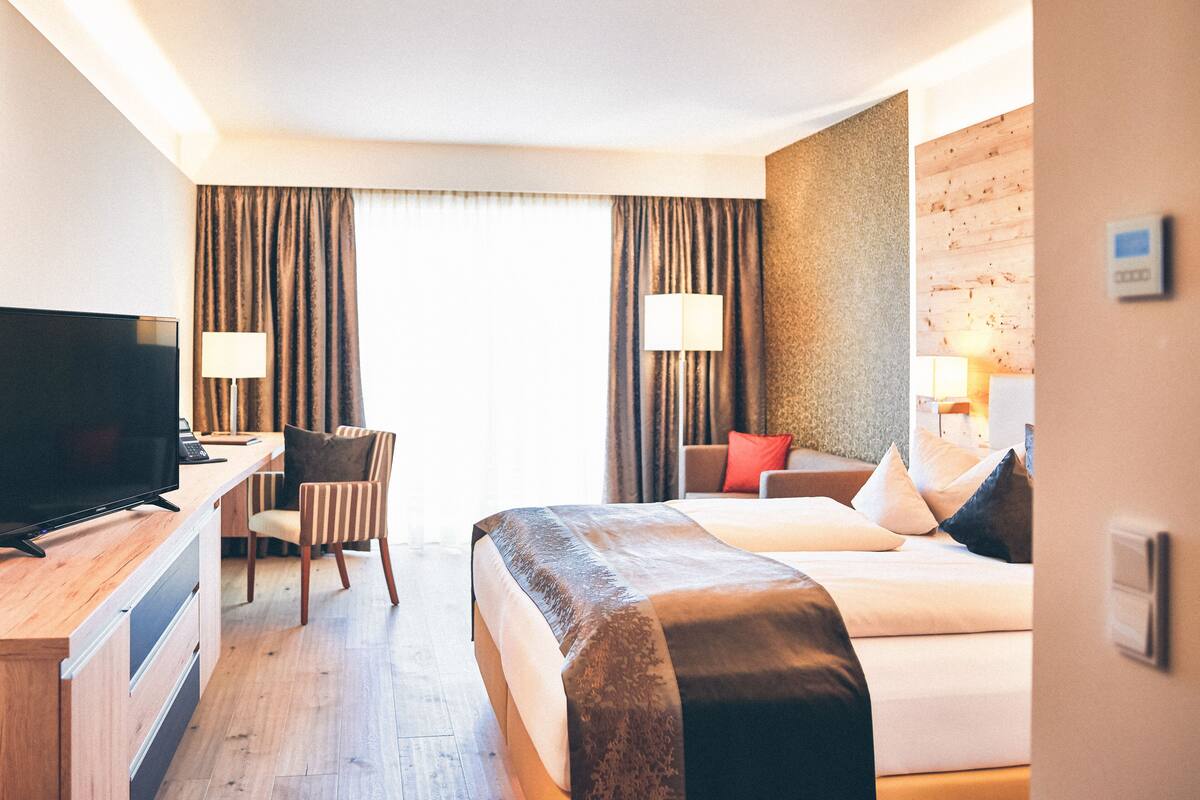 A hotel room with a bed, a desk with a TV, a chair, a lamp, and a view of the outdoors.
