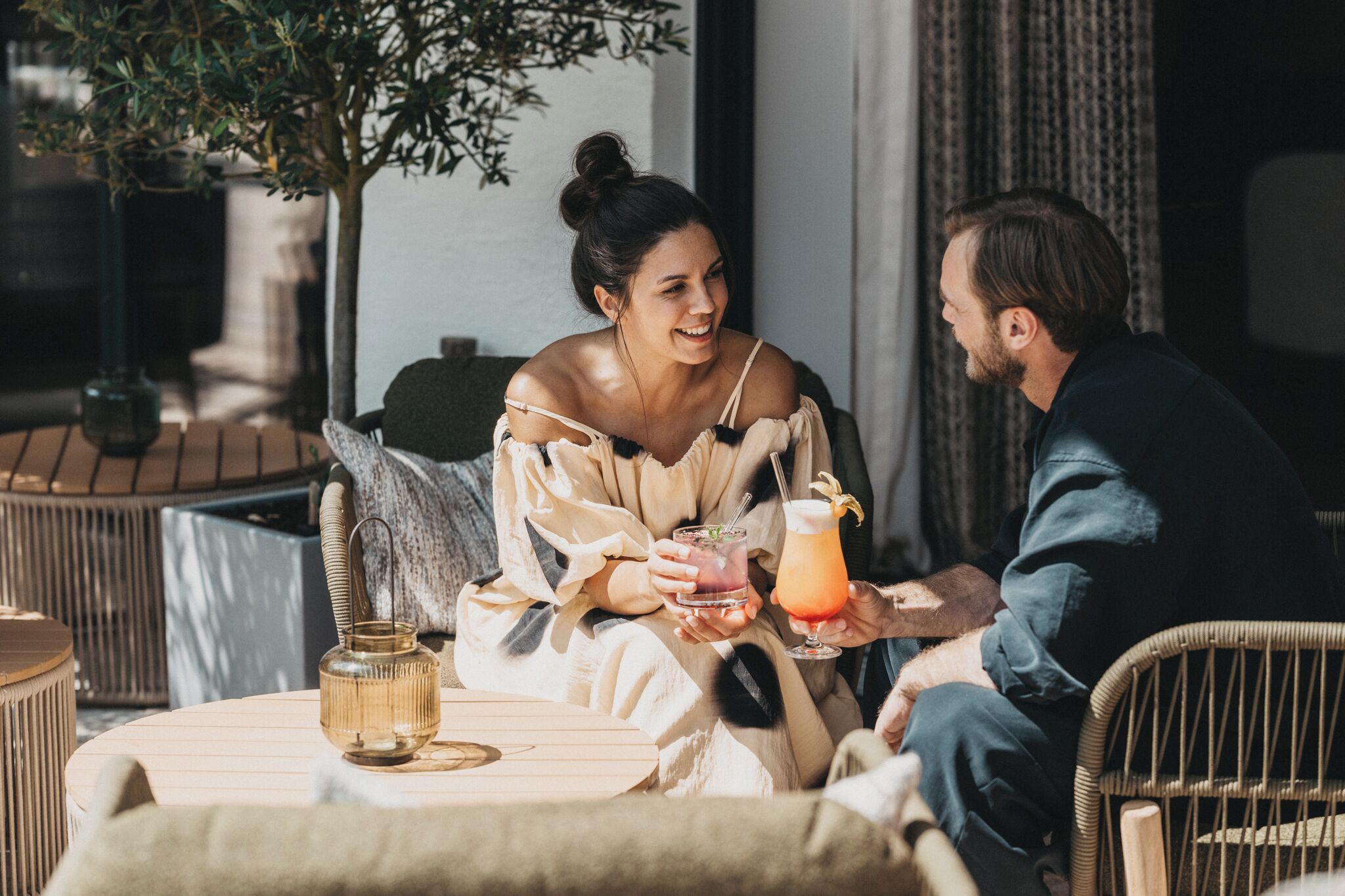 Two people seated at an outdoor table with drinks, surrounded by wicker furniture and a potted plant.