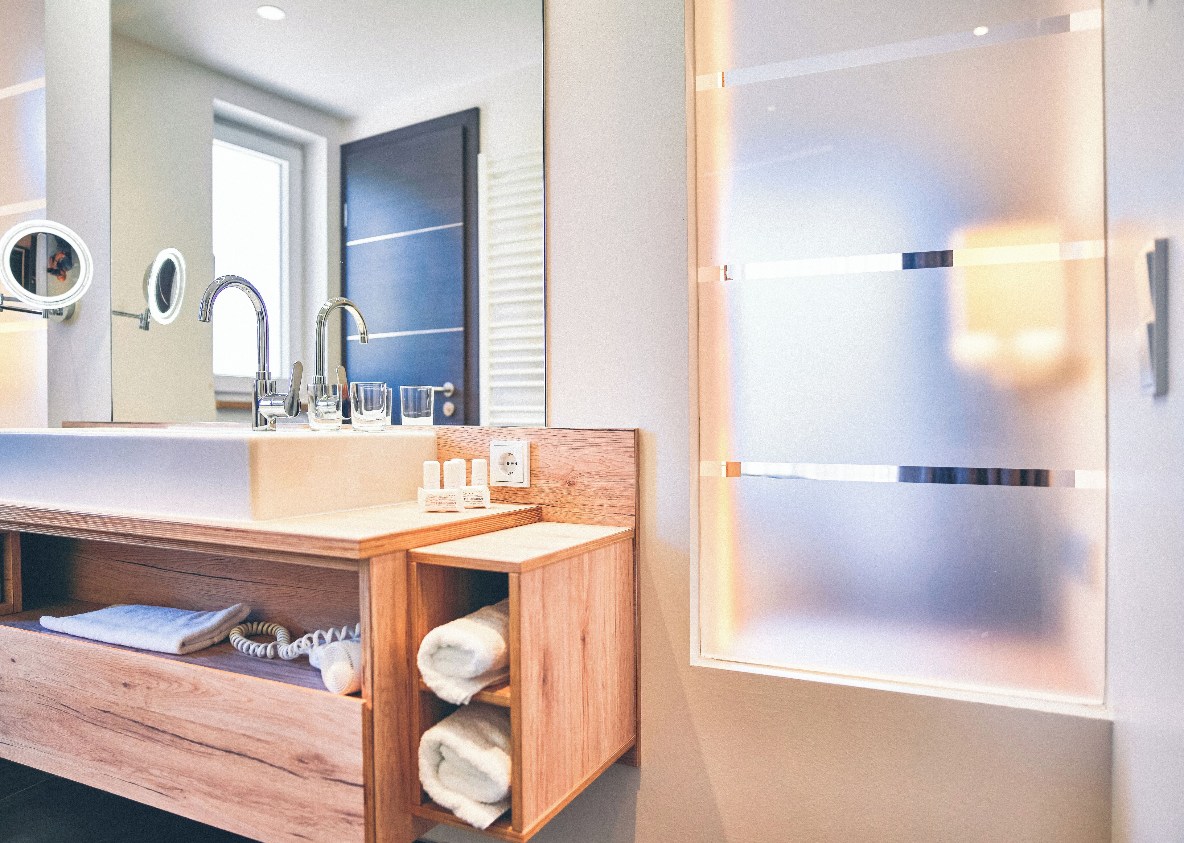 A modern bathroom with a wooden vanity, a large mirror, a sink, and a frosted glass cabinet.
