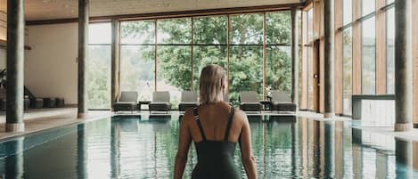 A woman sitting by an indoor pool with large windows and lounge chairs.