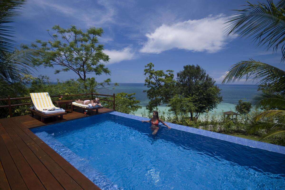 A pool area with a wooden deck, lounge chairs, and a view of the ocean.