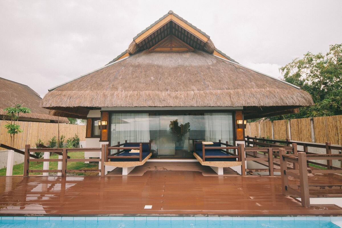 A thatched-roof building with a pool in front, wooden deck, and two seating areas.