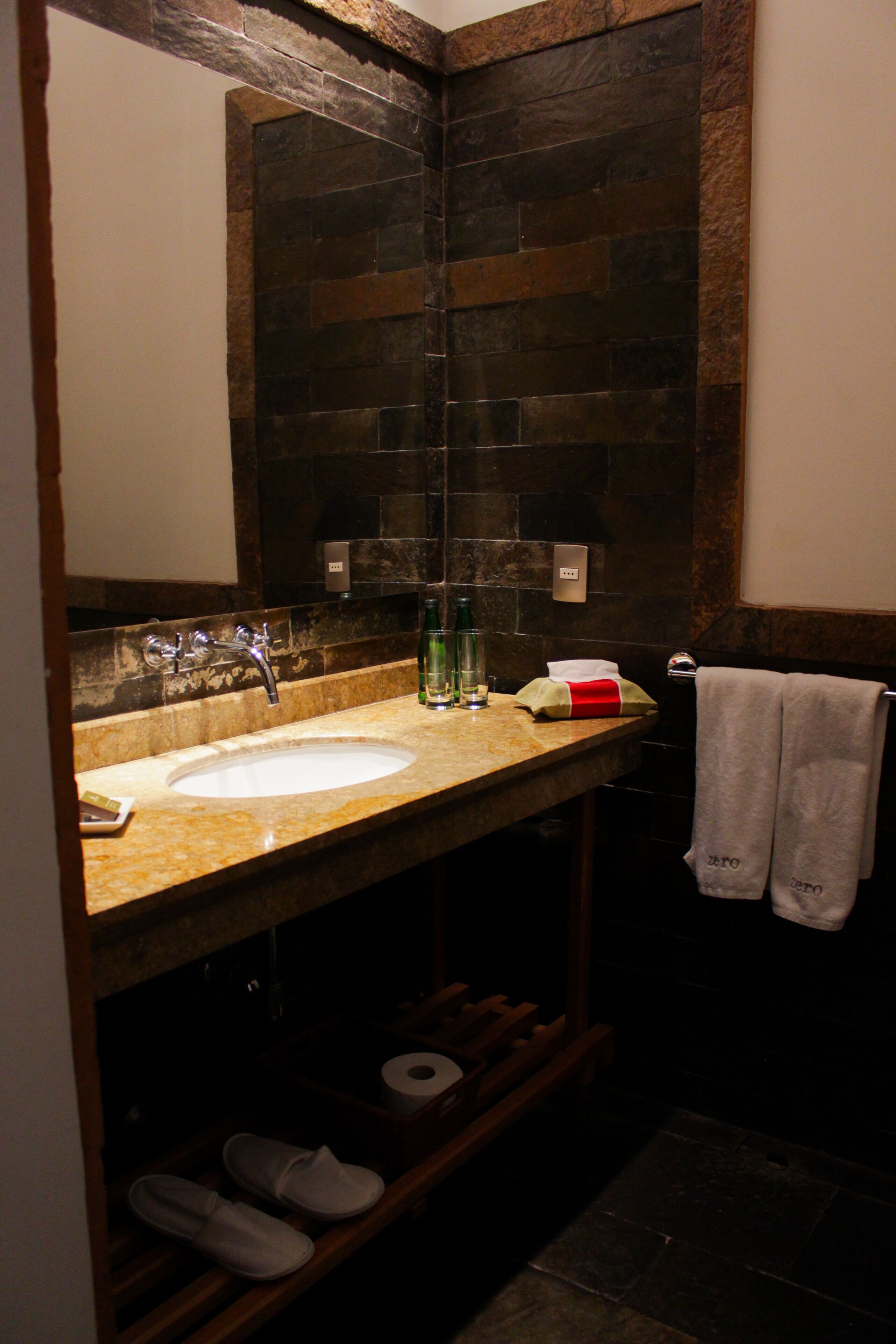 Une salle de bain avec un pare-feu en pierre, un lavabo, un miroir, un porte-serviettes et une étagère où sont rangées des chaussures.