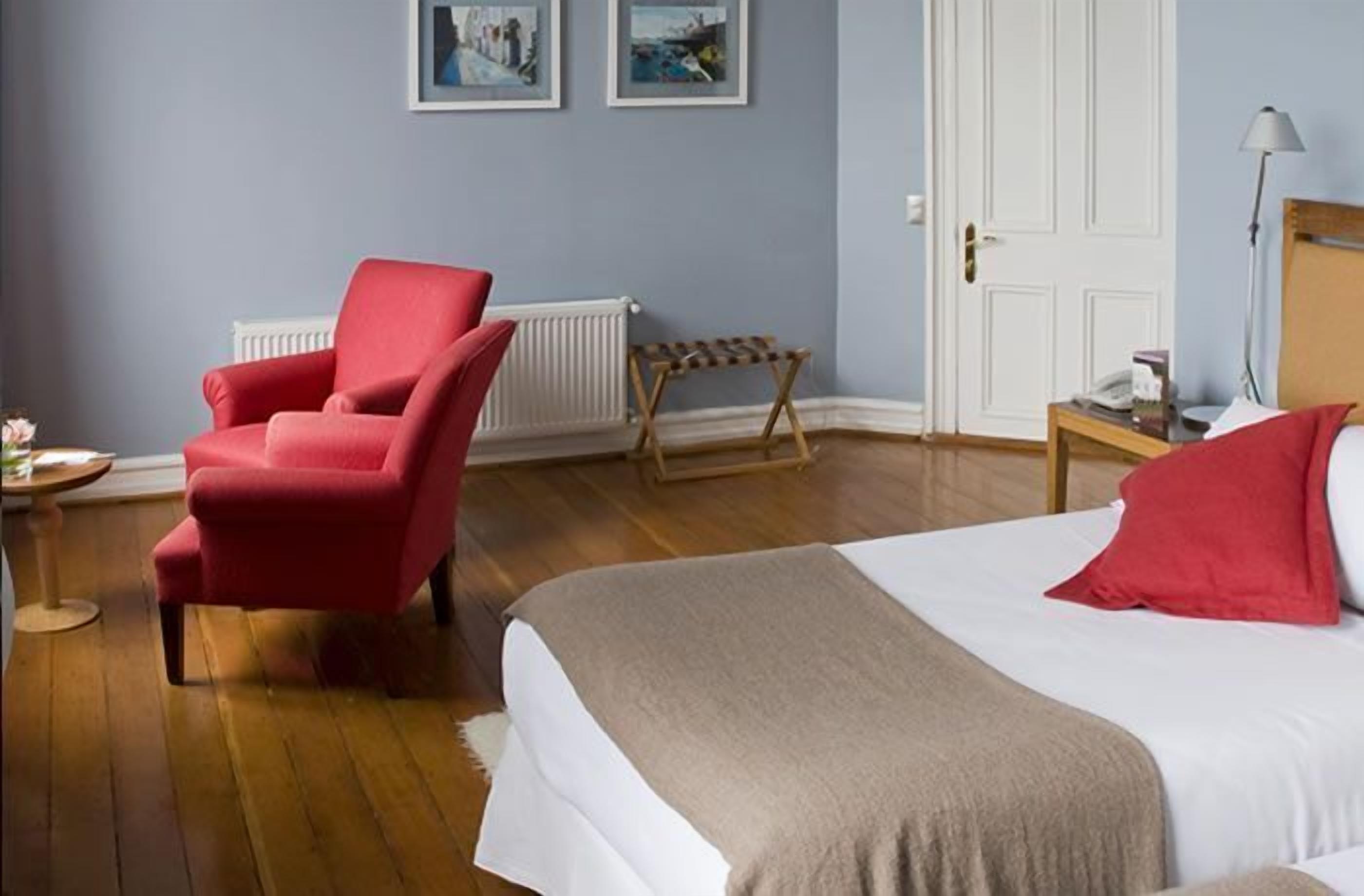 A bedroom with a bed, a red armchair, a wooden side table, and a radiator.