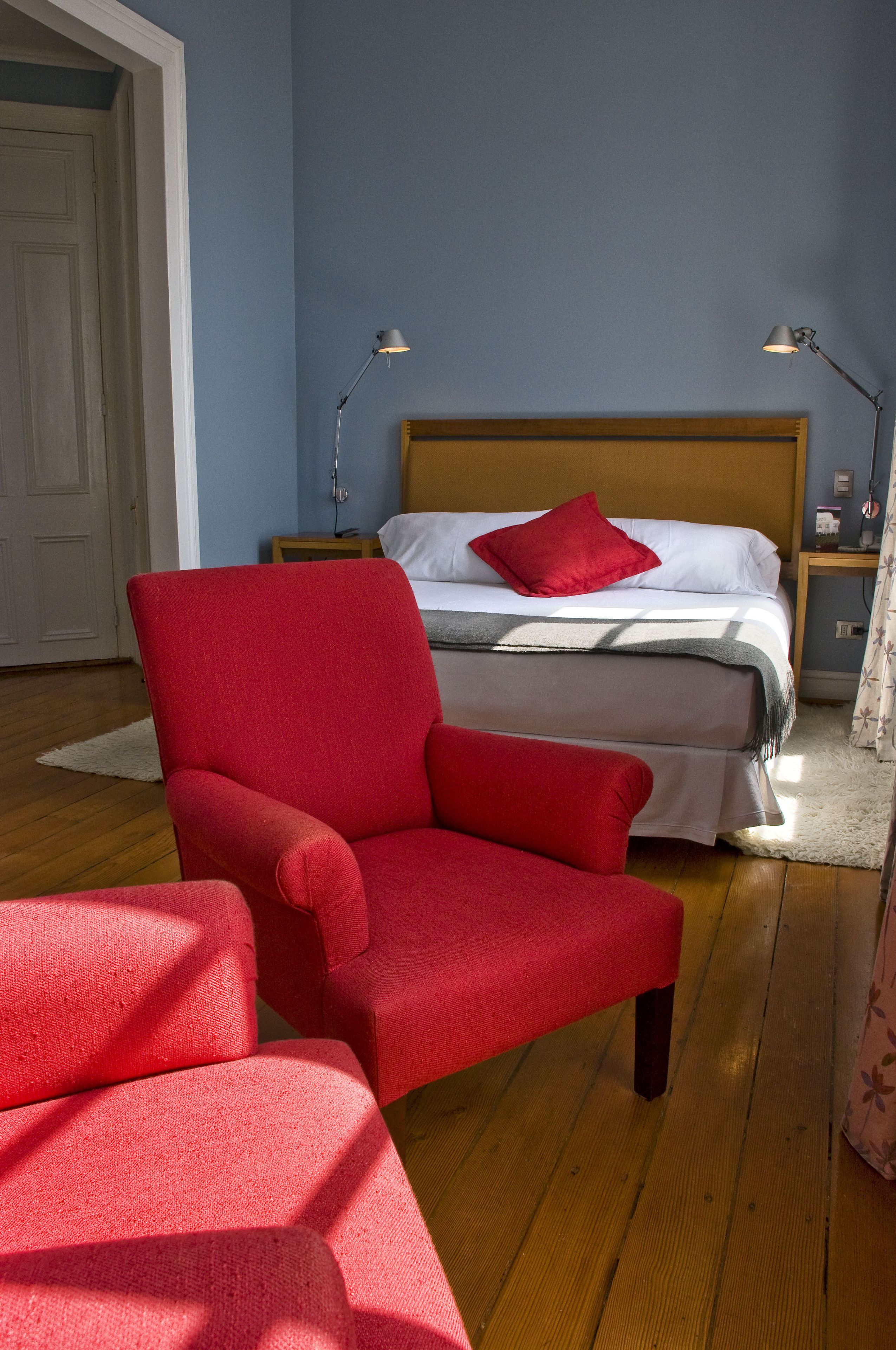A bedroom with a bed, two bedside lamps, a red armchair, and a wooden floor.