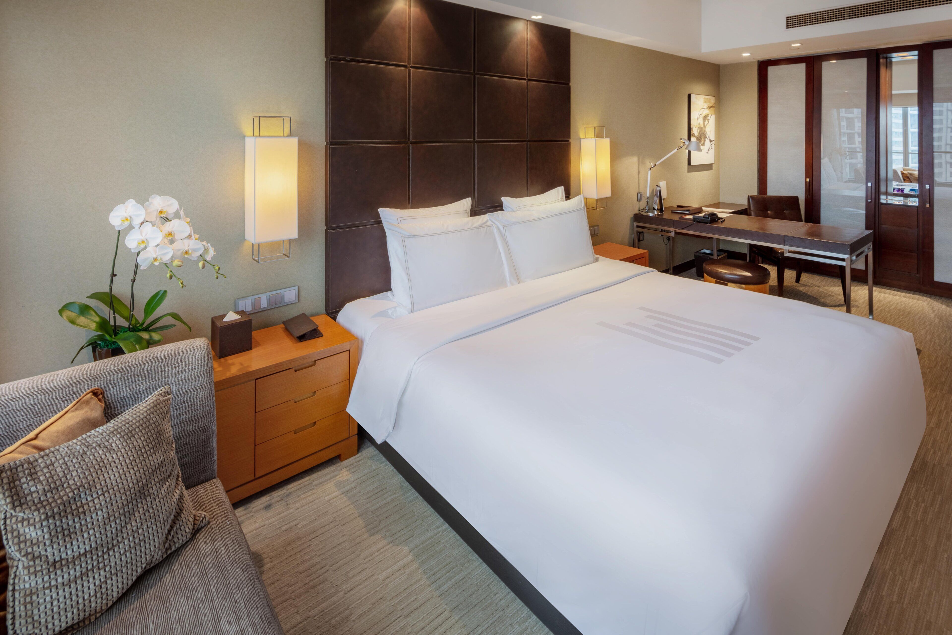 A hotel room with a large bed, a desk, a chair, a nightstand, a lamp, and a potted plant.