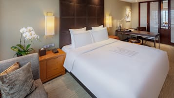 A hotel room with a large bed, a desk, a chair, a nightstand, a lamp, and a potted plant.