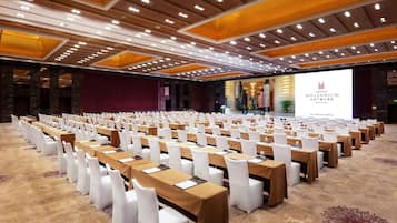 Un salón de conferencias con filas de mesas y sillas, un escenario con una pantalla y suelo alfombrado.