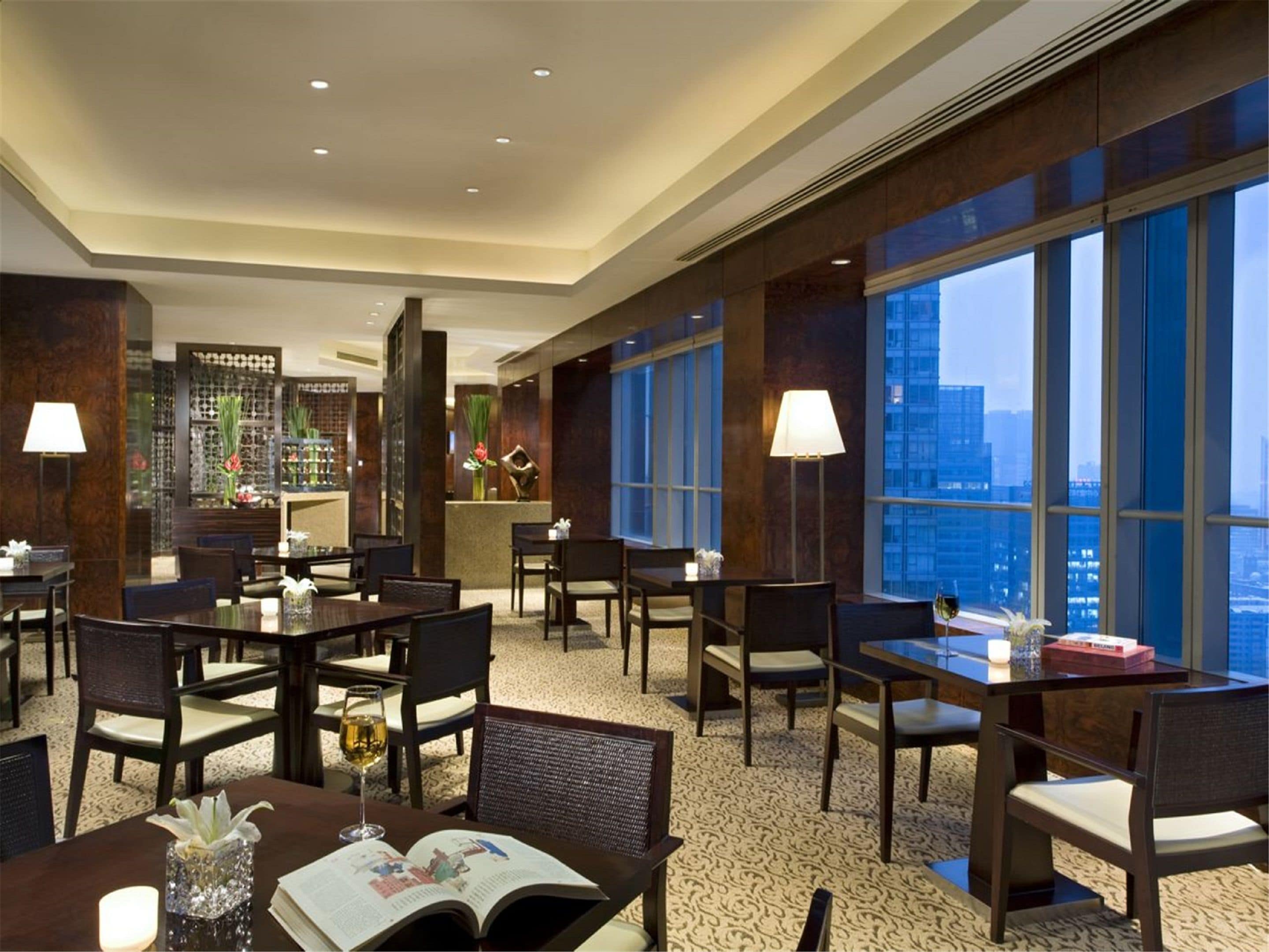 A modern hotel lobby with multiple dining tables and chairs, a reception desk, and large windows offering a city view.