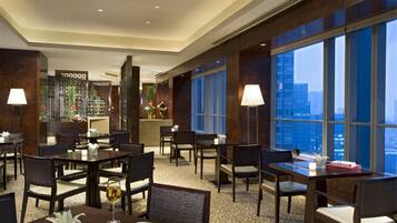 A modern hotel lobby with multiple dining tables and chairs, a reception desk, and large windows offering a city view.