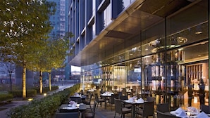 An outdoor dining area with tables and chairs, surrounded by glass walls and a view of the cityscape. - Grand Millennium Beijing (Beijing)