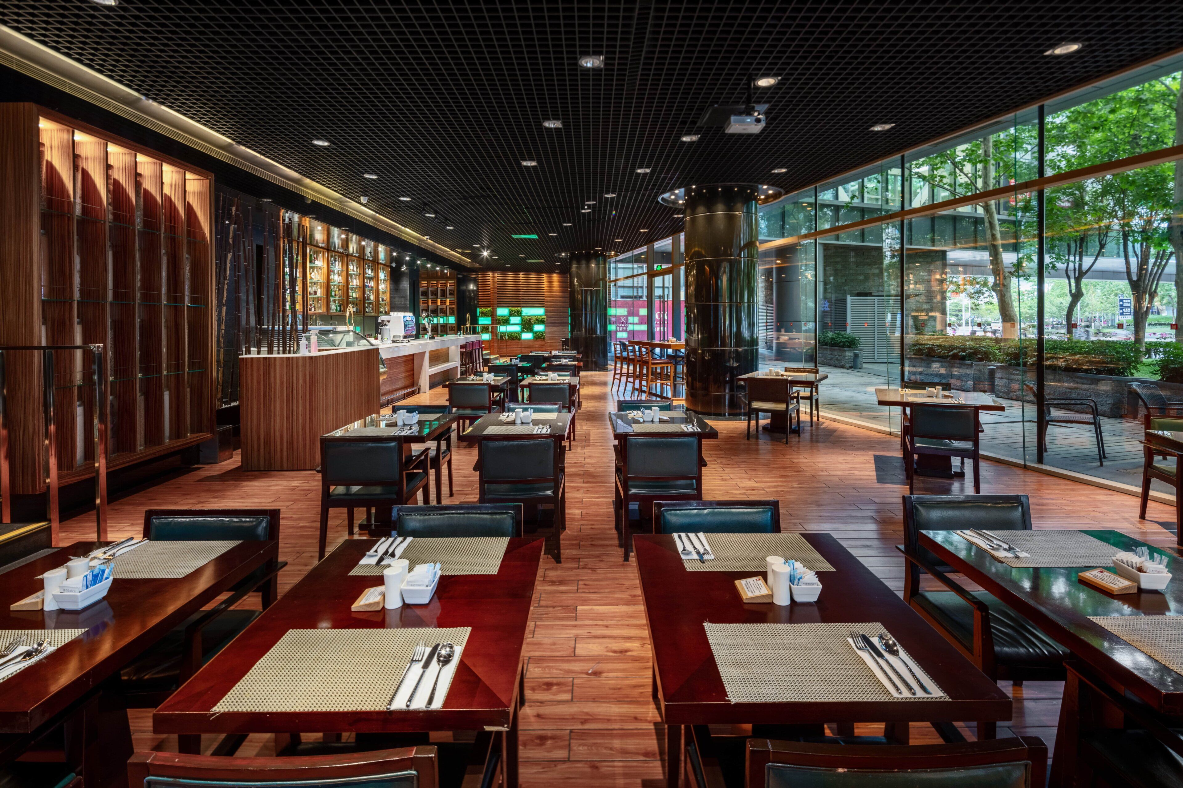 A modern restaurant with wooden tables and chairs, a bar area, and large windows offering a view of the outdoors.