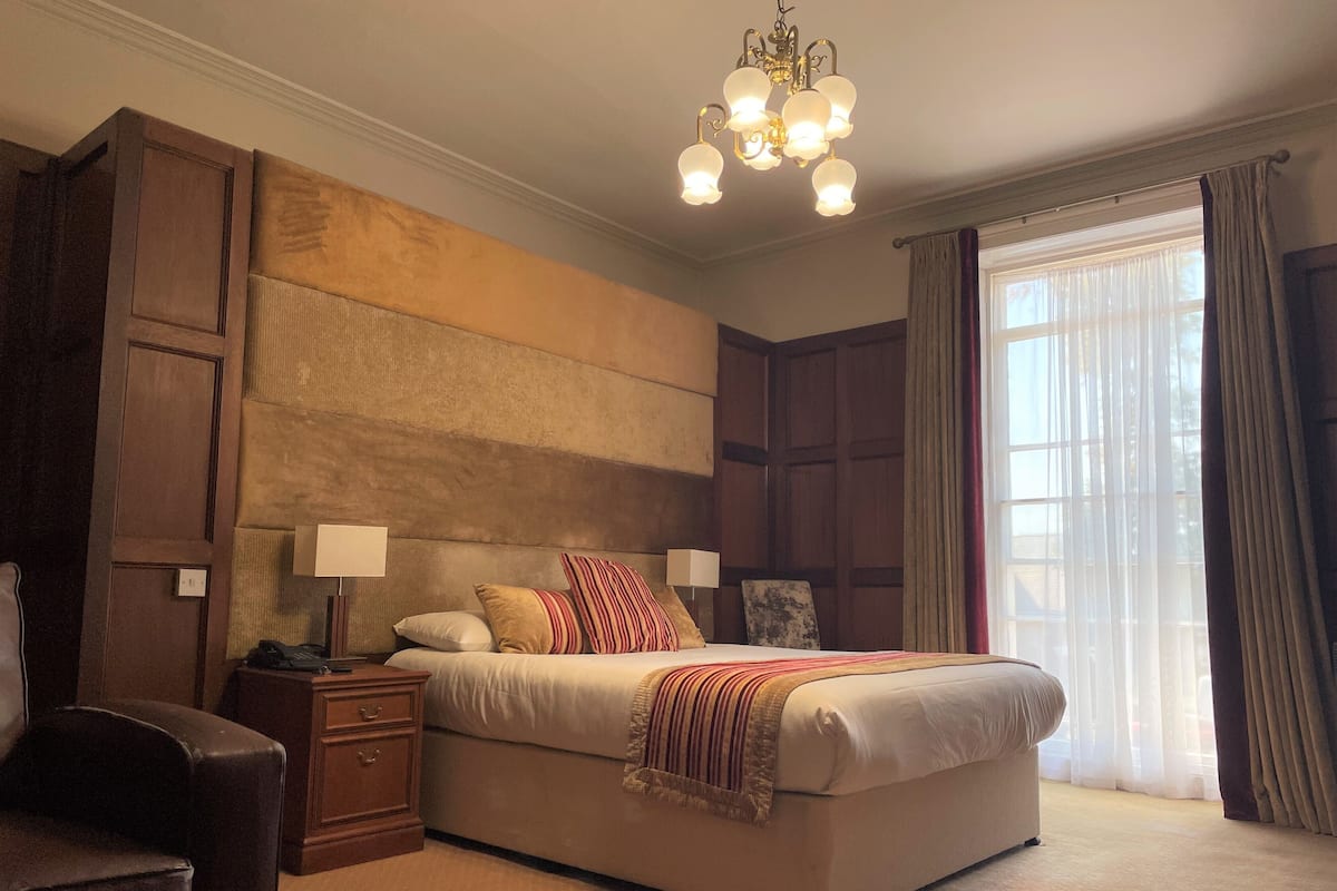 A well-lit bedroom with a large bed, wooden headboard, and a chandelier. There is a nightstand with a lamp, a chair, and a window with curtains.