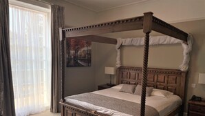 Ein Bett mit Baldachin, dessen Gestell aus Holz besteht und das ein wandpaneeliertes Kopfteil hat. Ein Nachttisch mit Lampe steht daneben, und an der Wand hängt ein Gemälde.